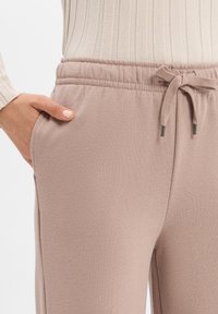 Ręka opiera się w kieszeni spodni dresowych o podwyższonym stanie w odcieniu różowego brzoskwini, związywanych w pasie sznurkiem, w zestawieniu z beżowym, prążkowanym topem z długim rękawem.