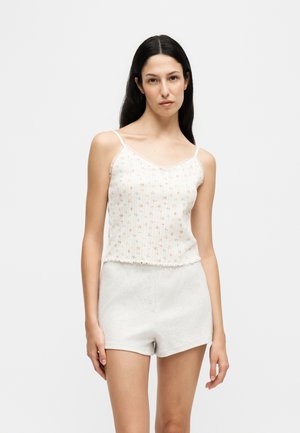Femme aux longs cheveux foncés portant un haut blanc sans manches à motifs floraux et un short gris clair sur un fond uni.