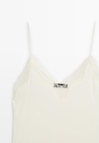 Cami-top in crèmekleur met een kanten rand rond de halslijn en verstelbare spaghettibandjes; zachte stof; etiket zichtbaar in het midden.