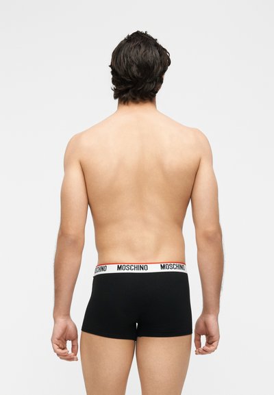 Άνδρας μοντέλο στέκεται με την πλάτη στην κάμερα, φοράει μαύρα boxer briefs Moschino με λευκή ζώνη με λογότυπο.