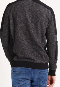Pull gris avec des accents noirs, texture tricotée, encolure ronde et poignets côtelés. Porté avec un jean en denim bleu.