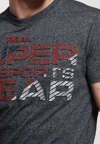 Šedé tričko s krátkým rukávem s výrazným, barevným nápisem "SKUTEČNÉ SUPERSPORTOVNÍ VYBAVENÍ", s texturou písma v červené, bílé barvě a pruhovanými vzory.