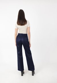 Granatowe spodnie o wysokim stanie z gładką fakturą i szerokimi nogawkami. Połączone z dopasowaną beżową bluzką, na białym tle.