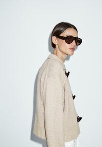 Beige knitted sweater with a collar, featuring small brown bow accents. Model wearing dark, oversized sunglasses against a white background.