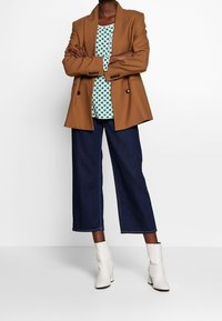 Brauner maßgeschneiderter Blazer über einem grünen gepunkteten Oberteil, kombiniert mit weit geschnittenen, dunklen Jeans und weißen Knöchelstiefeln für einen geschichteten Look.