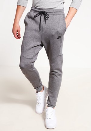 Personne portant un pantalon de survêtement gris Nike avec un cordon noir et des baskets blanches, debout sur fond blanc.