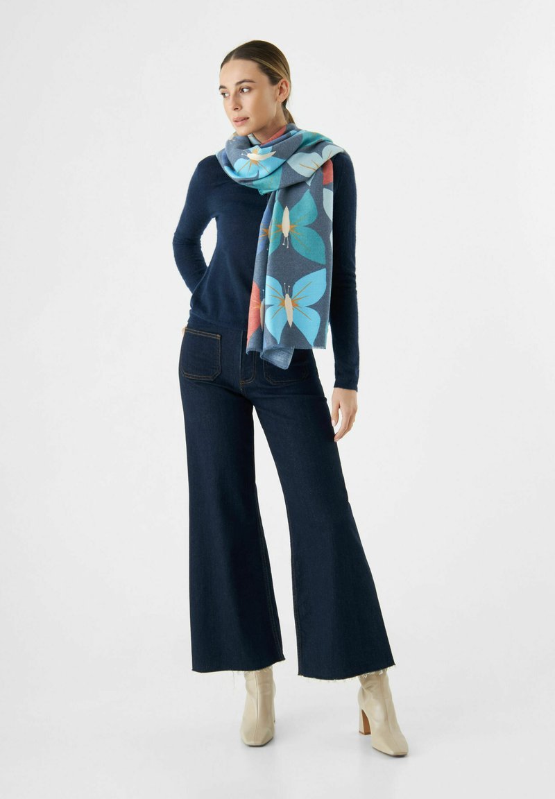 Un pull en tricot bleu, un jean foncé évasé et une écharpe bleue à motifs de papillons aux couleurs vives, portés avec des bottines beige clair.