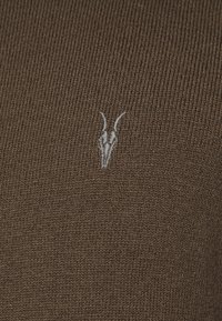 Hnědá pletená látka s jemným vyšívaným motivem beraní hlavy v světle šedé barvě, který přidává oděvu jedinečný detail.