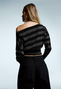 Fekete és fémes csíkos, rövid fazonú pulóver vállakat szabadon hagyó kialakítással és fodros ujjakkal, fekete magas derekú farmerrel párosítva.