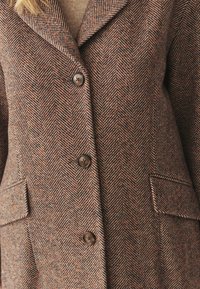 Blazer de tweed en patrón de espiga marrón y naranja, con solapas de muesca, cuatro botones y dos bolsillos delanteros.