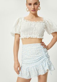 Hvid cropped top med tekstureret blonde, puffede ærmer og bølget kant. Lyseblå nederdel med blomstermotiv, rynket side og volangkant.