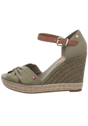 Beżowa sandała espadrylowa na koturnie z tkanym obcasem, z przodu zawiązana na supeł z tkaniny, brązowym paskiem wokół kostki oraz małym czerwono-biało-niebieskim logo po boku.