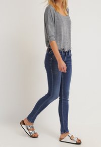 Blusa gris de manga larga y corte holgado, jeans ajustados de color azul oscuro y sandalias blancas con hebillas plateadas. Suela plana y texturizada, vista lateral.