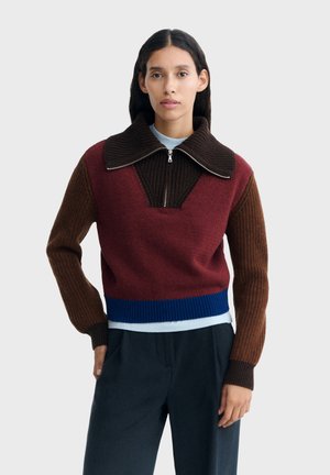 Donna che indossa un maglione color-block con corpo color bordeaux, maniche marroni, collo marrone scuro con cerniera e orlo blu, abbinato a pantaloni neri.