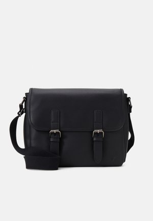 Sac messager en cuir noir avec rabat, deux sangles à boucles sur le devant et bandoulière en tissu réglable sur fond blanc.