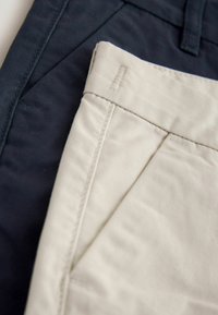 Deux pantalons pliés ensemble : un bleu marine, un beige. Les deux sont en coton lisse avec des coutures droites et des détails de surpiqûres subtils.