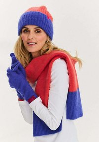 Pletená čepice v růžové a modré barvě, ladící modrý šátek a rukavice, vyrobené z měkkého, texturovaného materiálu. Na rukavicích je jemná značka značky.