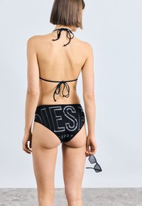 Μαύρο μπικίνι με halter top και κλείσιμο με φιόγκο, καθώς και ψηλόμεσα σλιπ που εμφανίζουν έναν τολμηρό λογότυπο. Λείο υλικό, μινιμαλιστικός σχεδιασμός.