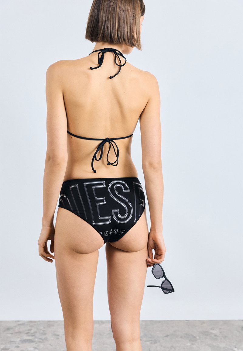 Μαύρο μπικίνι με halter top και κλείσιμο με φιόγκο, καθώς και ψηλόμεσα σλιπ που εμφανίζουν έναν τολμηρό λογότυπο. Λείο υλικό, μινιμαλιστικός σχεδιασμός.