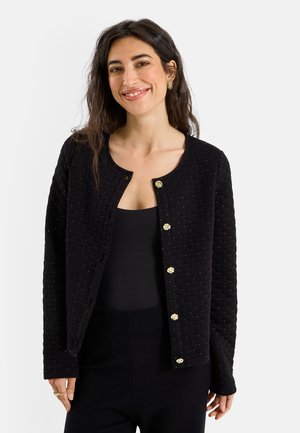 Mujer con el cabello largo y oscuro, llevando una chaqueta negra acolchada con botones dorados sobre una camiseta negra, sonriendo sobre un fondo blanco.