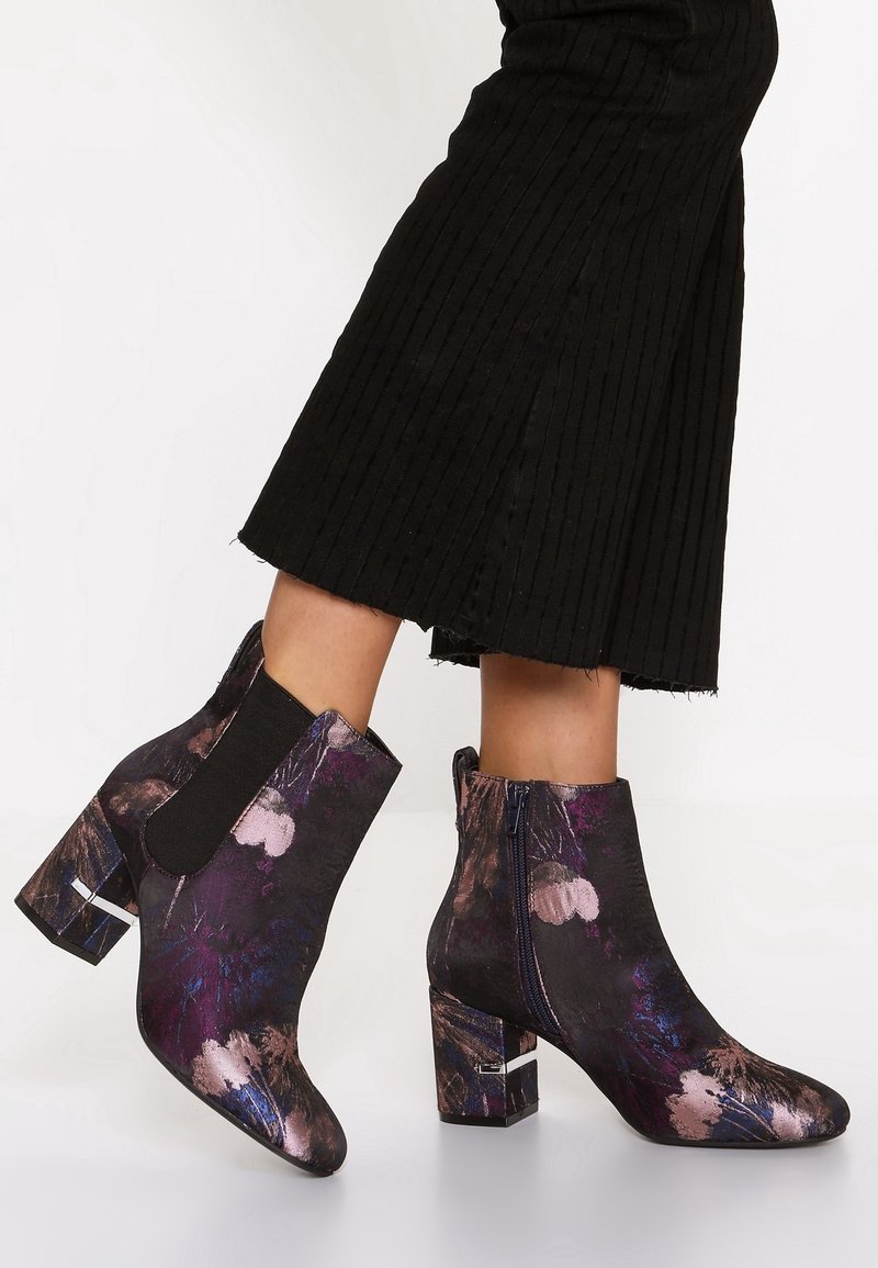 Bottes à talon bloc hauteur cheville avec un motif floral violet et noir, portées avec une jupe noire côtelée.