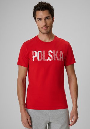 Mężczyzna z krótkimi kręconymi włosami ubrany w czerwoną koszulkę z napisem "Polska" białymi literami i jasnoszare spodnie na jednolitym tle.