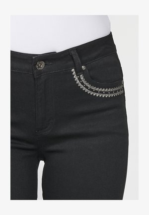 Schwarze Jeans mit einer Vordertasche, verziert mit silbernem dekorativem Stick und Verzierungen, einem Knopfverschluss und standardmäßiger Denim-Textur.
