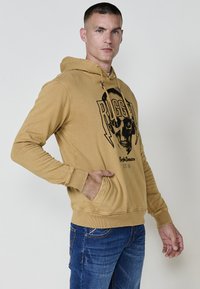 Bežs kapuce džemperis ar melnu skeleta grafiku un tekstu "RUGGED", aprīkots ar kanguruma kabatu un ribinātām piedurknēm, izgatavots no mīkstas, teksturētas auduma.