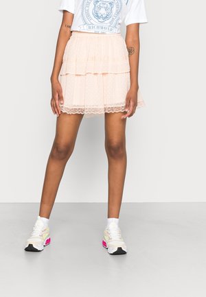 Jeune personne portant un t-shirt graphique blanc, une mini-jupe en dentelle rose pâle superposée, des chaussettes blanches et des baskets blanches avec des accents roses.