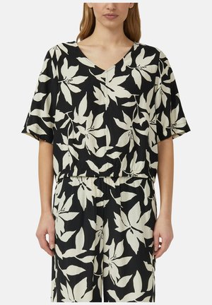 Femme portant un haut à manches courtes imprimé floral noir et blanc et un pantalon large assorti avec une taille élastique, debout devant un fond uni.