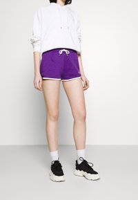 Shorts en coton violet avec des bordures blanches, associés à un hoodie blanc et des baskets noires avec des semelles blanches. Détail de cordon de serrage simple à la taille.