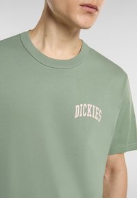 Zelené bavlněné tričko s krátkým rukávem, s zakřiveným růžovým textem loga "DICKIES" na levé části hrudníku. Hladká textura a klasický střih.