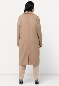 Cappotto lungo in lana beige con colletto semplice, spacco posteriore e maniche raglan, abbinato a pantaloni beige chiaro e tacchi blu.