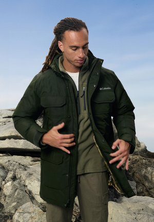 Chaqueta acolchada verde con múltiples bolsillos frontales y un collar alto. Debajo, una sudadera con cremallera gris. Fondo neutro con rocas.