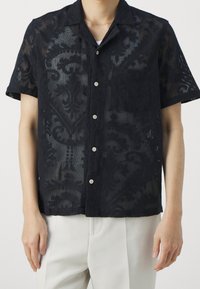 Camicia nera a maniche corte realizzata in tessuto semi-trasparente con un intricato motivo tonale. Dettagli notevoli includono una tasca e una chiusura con bottoni.