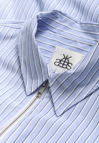 Lyseblå skjorte med hvide striber, der har en spids krave og lynlås foran. Stoffet ser glat og let tekstureret ud. Mærkat synligt.