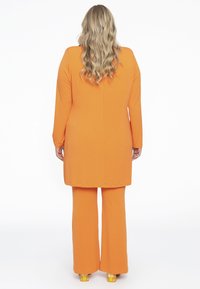 Orange Langarm-Tunika und passende, weit geschnittene Hosen aus glattem Stoff, mit geradem Schnitt, sichtbar sind klobige, gemusterte Absätze.