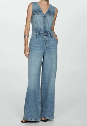 Combinaison en denim sans manches avec boutons sur le devant, pantalons larges, poches poitrine et passants de ceinture, portée avec des bracelets et des chaussures noires à bout ouvert.