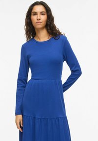 Modré dlouhé šaty s dlouhým rukávem v žebrované textuře s přiléhavým živůtkem a vrstvenou sukní. Mají kulatý výstřih a hladký povrch tkaniny.