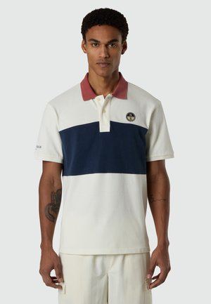 Mann trägt ein kurzärmeliges Polohemd mit rostfarbenem Kragen, blauem Streifen auf der Brust und beige unterem Bereich, steht vor einem einfarbigen Hintergrund.