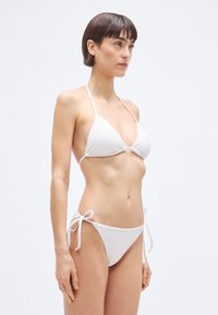Σετ λευκό μπικίνι που περιλαμβάνει τριγωνικό τοπ με ελάχιστη επένδυση και εσώρουχα με ρυθμιζόμενο δέσιμο στο πλάι. Ομαλή υφή και απλός σχεδιασμός.