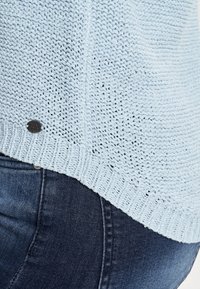 Lichtblauwe gebreide trui met een structuurpatroon en geribde zoom; voorzien van een klein metalen logo-label aan de zijkant, gecombineerd met donkerblauwe jeans.