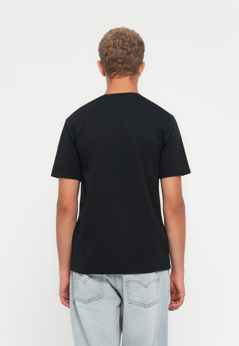 Osoba z krótkimi kręconymi włosami, ubrana w gładką czarną koszulkę z krótkim rękawem i jasnoniebieskie dżinsy, stoi tyłem na białym tle.