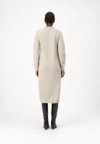 Robe tricot beige à manches longues avec un col rond et des poignets côtelés, tombant en dessous des genoux, assortie à des bottes noires à genoux.