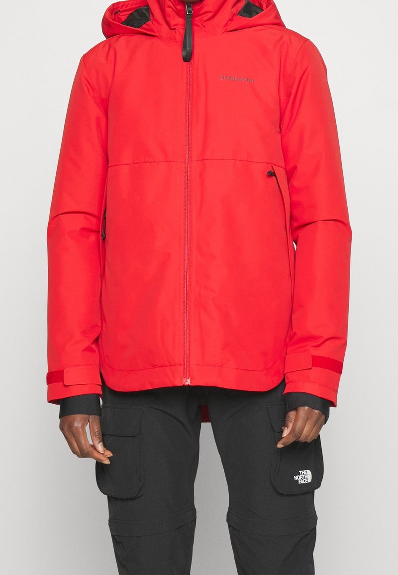 Homme portant une veste à capuche rouge vif avec fermeture éclair et un pantalon noir avec poches latérales et le logo The North Face.