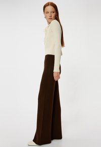 Crème geribbelde trui met een voorknoop, gecombineerd met hoge bruine broek met wijde pijpen. Gelaagd materiaal, strakke lijnen en een getailleerd silhouet.