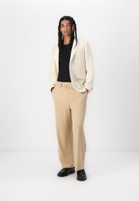 Lichtbeige blazer over een zwarte top, gecombineerd met losse beige broeken. De outfit heeft strakke lijnen en een relaxte pasvorm, met zwarte schoenen.