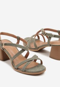 Sandales à talons en tissu vert avec des lanières entrecroisées, des accents tan et des talons en blocs texturés. Design à bout ouvert avec une semelle en caoutchouc.