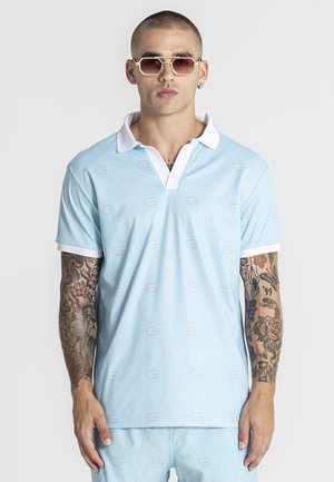 Uomo con capelli corti e tatuaggi sulle braccia che indossa una polo azzurro chiaro, pantaloncini abbinati e occhiali da sole esagonali, in piedi davanti a uno sfondo bianco.