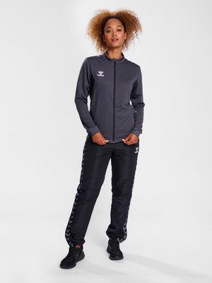 Kvinde med krøllet hår står iført en mørkegrå sportjakke med lynlås og sorte træningsbukser med sidestriber samt sorte sneakers.